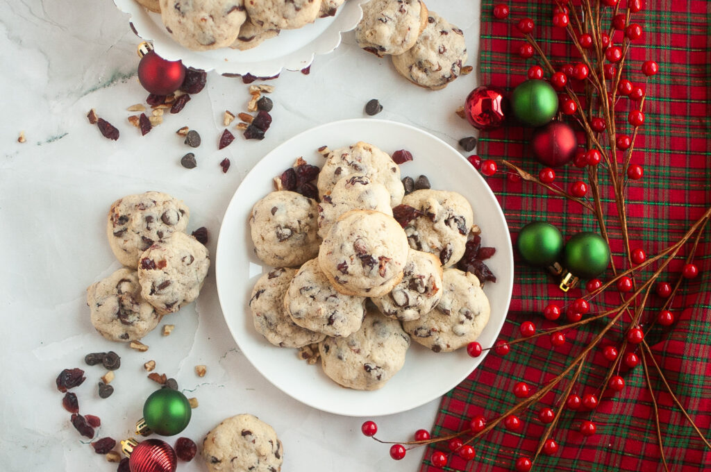 A plate and stand of chocolate chip cookies with cranberries and walnuts, surrounded by red and green ornaments and red berries on a plaid holiday cloth.