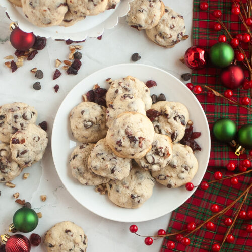 A plate and stand of chocolate chip cookies with cranberries and walnuts, surrounded by red and green ornaments and red berries on a plaid holiday cloth.