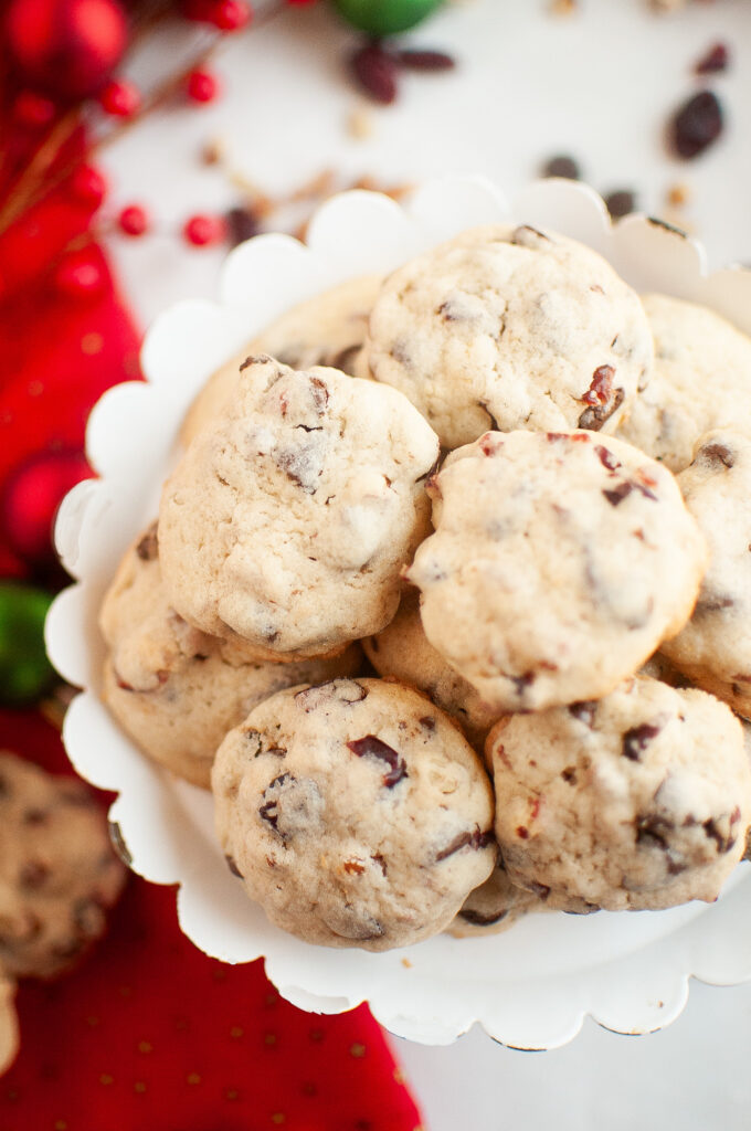 A white plate holds a pile of chocolate chip cookies with visible pieces of chocolate and cranberries. A red cloth and holiday decorations are partially visible in the background.