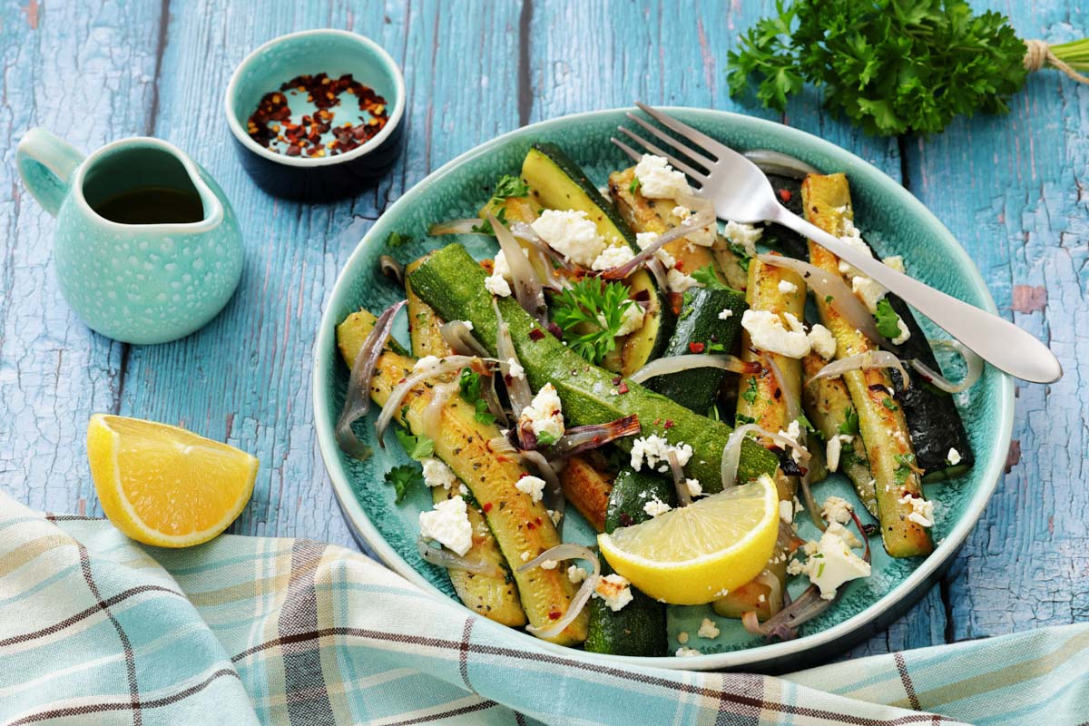 A plate of grilled zucchini with onions, crumbled cheese, and herbs, garnished with lemon wedges, set on a blue wooden table with a fork, napkin, oil jug, and bowl of spices nearby.
