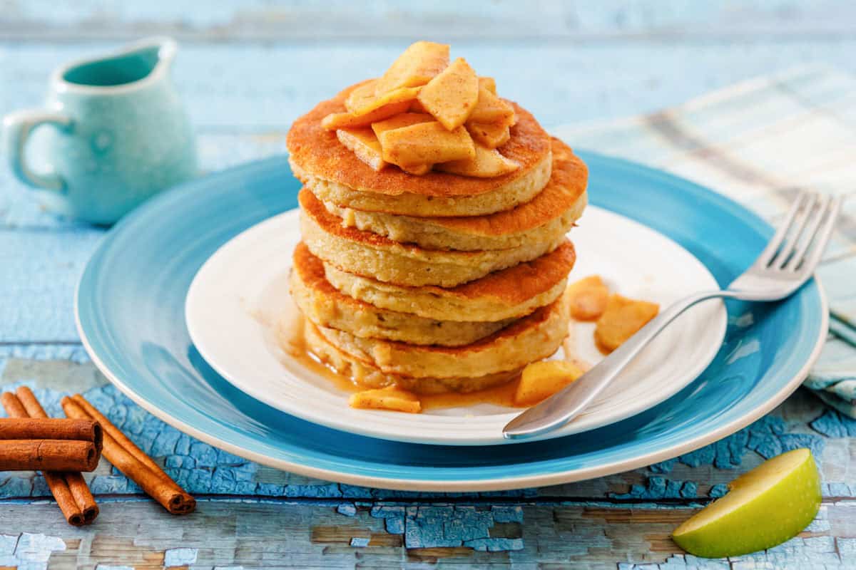 A stack of pancakes topped with cooked apple slices and syrup on a white plate with a blue rim, next to a fork, cinnamon sticks, and a small jug.