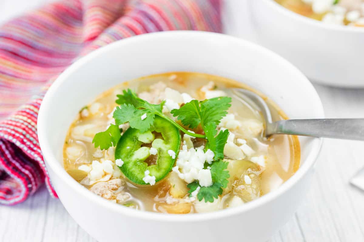 A bowl of Green pork posole soup garnished with cilantro, sliced jalape&ntilde;o, and crumbled cheese, with a spoon resting inside and a striped cloth in the background.