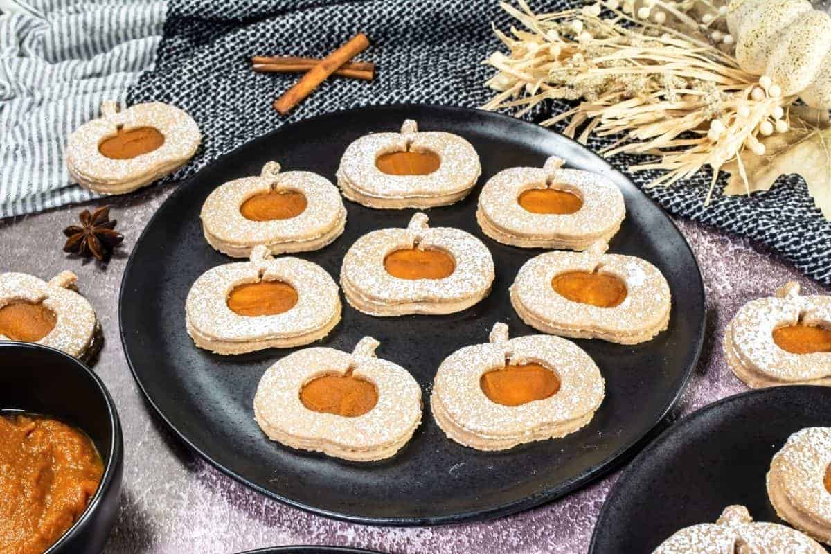 Plate of pumpkin-shaped cookies with powdered sugar and pumpkin filling, arranged on a black dish with decorative autumn items and spices in the background.