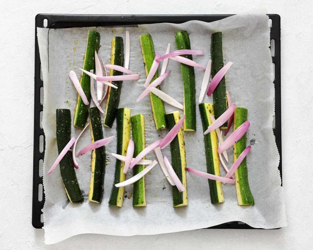 Zucchini sticks and sliced shallots arranged on a parchment-lined baking tray, ready for roasting.