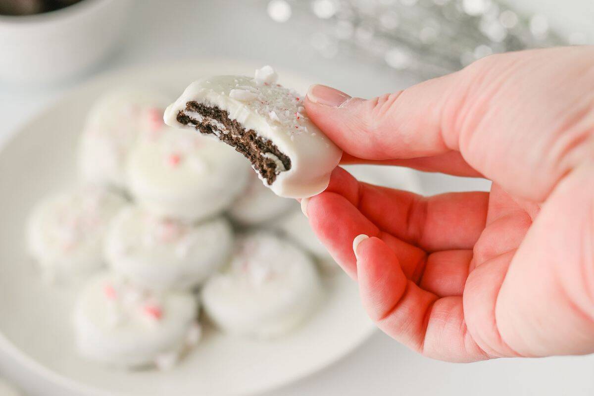 A hand holds a Peppermint Crunch Oreo with a bite taken out, with more cookies on a plate in the background.
