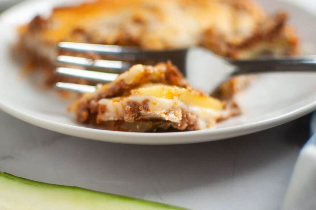 Close-up of a fork cutting into a slice of lasagna on a white plate, with layers of cheese, sauce, and pasta visible.