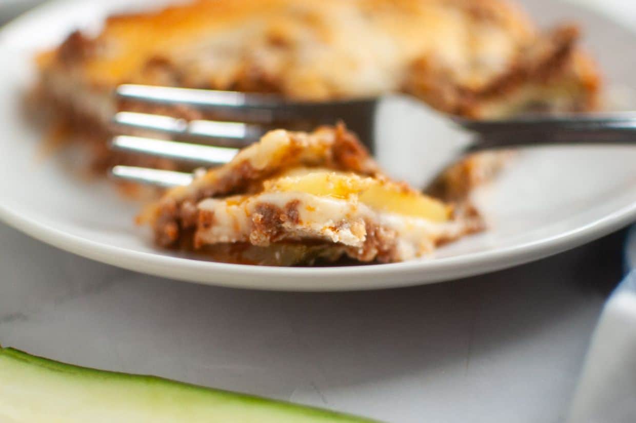 Close-up of a fork cutting into a slice of lasagna on a white plate, with layers of cheese, sauce, and pasta visible.
