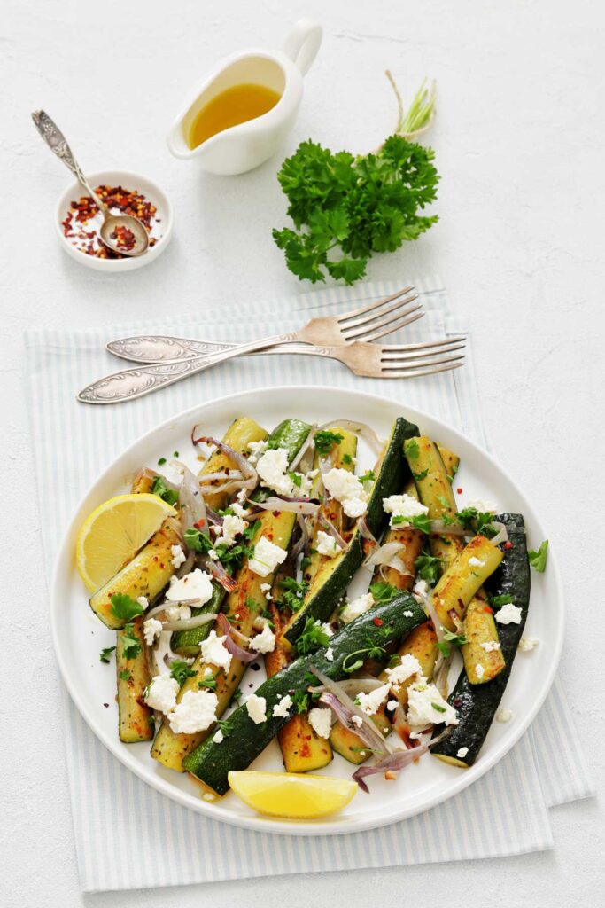 A plate of grilled zucchini with red onions, crumbled feta, parsley, lemon wedges, and a drizzle of olive oil, served with a side of chili flakes and fresh herbs on a table.