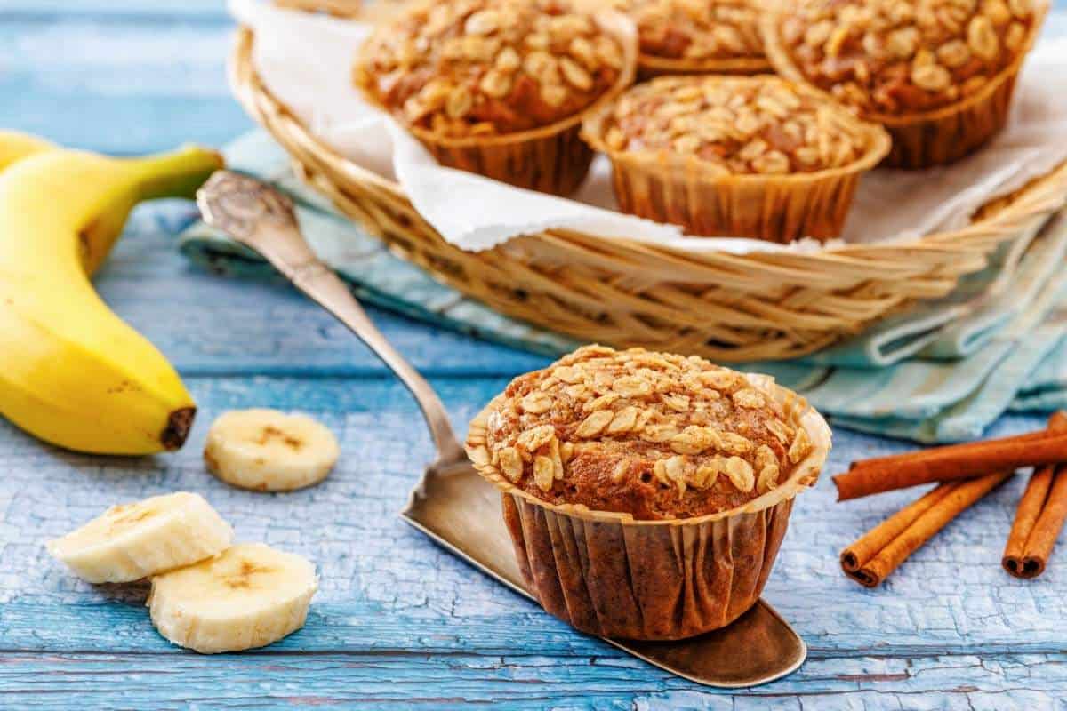 Banana oatmeal muffins on a metal spatula and in a basket, with sliced bananas and cinnamon sticks on a blue wooden surface.