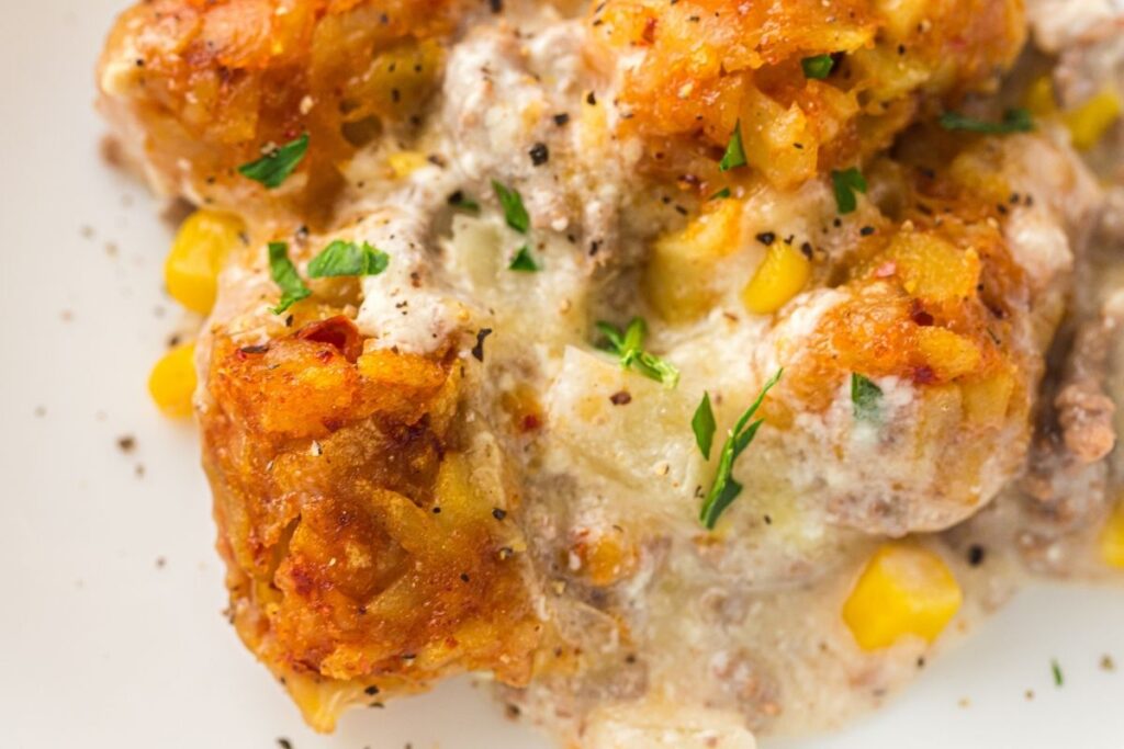 Close-up of a tater tot casserole with ground meat, corn, creamy sauce, melted cheese, and herbs, topped with ground black pepper.