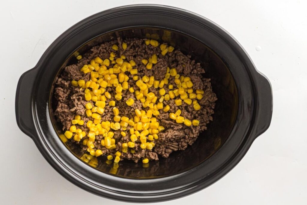 Ground beef and corn kernels are placed inside a black slow cooker on a white surface.