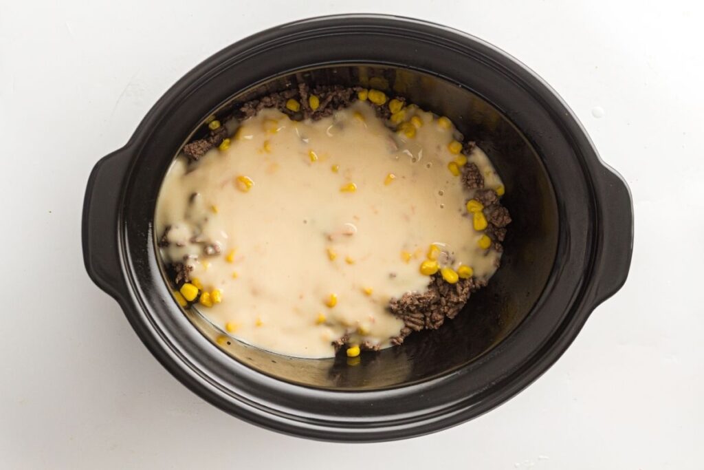 A slow cooker filled with ground beef, corn, and a creamy soup mixture, viewed from above on a white background.