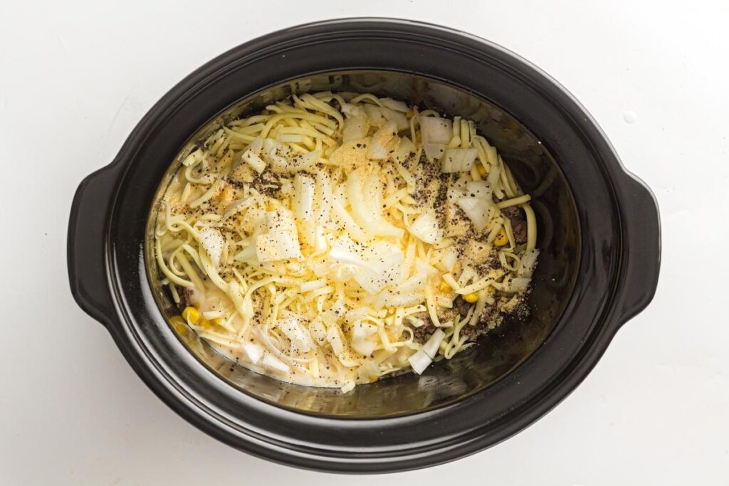 Uncooked cheddar, chopped onions, corn, seasonings, and liquid in a black slow cooker viewed from above.