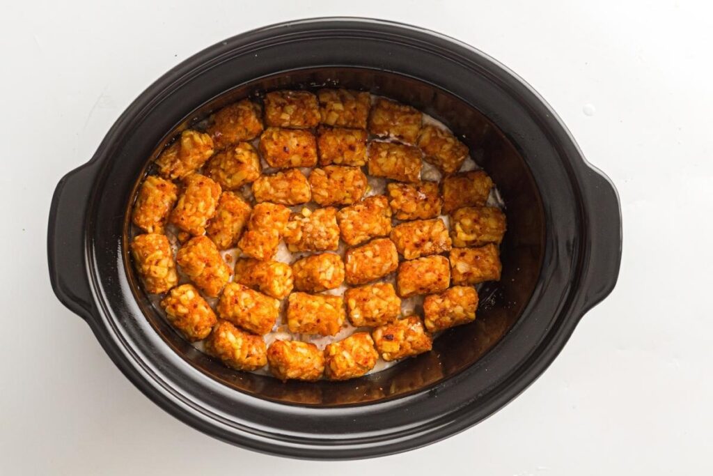 A black slow cooker filled with a single layer of tater tots arranged neatly on a creamy mixture.