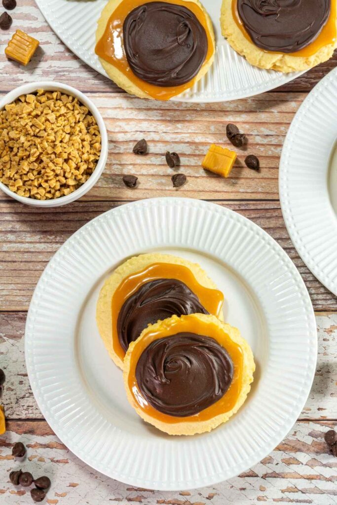 Two sugar cookies topped with caramel and chocolate are on a white plate, with a bowl of toffee bits, chocolate chips, and caramel pieces nearby on a wooden table.