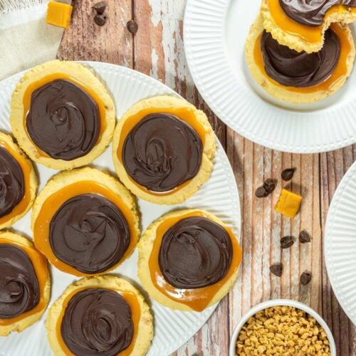 A plate of Copycat Crumbl Twix Cookies, with more cookies on separate plates, caramel pieces, and small chocolate chips nearby on a wooden table.