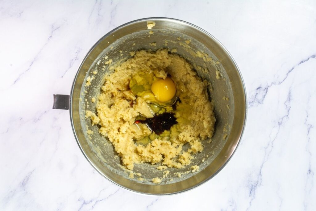 A mixing bowl with cookie dough, an egg, and vanilla extract on top, placed on a white marble surface.