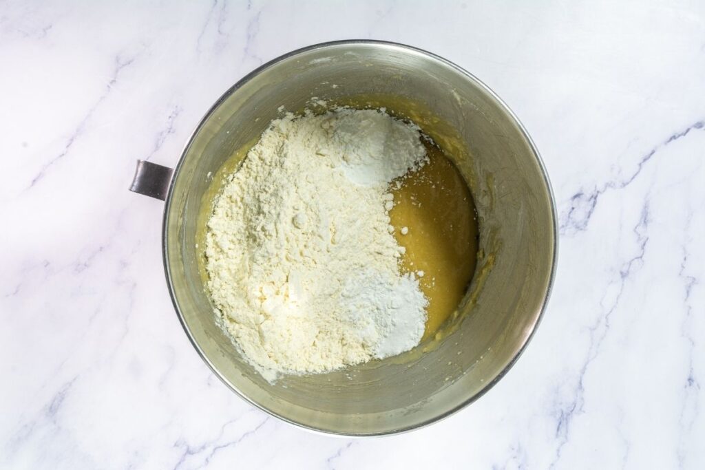 A metal mixing bowl with partially mixed dough and flour on top, placed on a white marble surface.