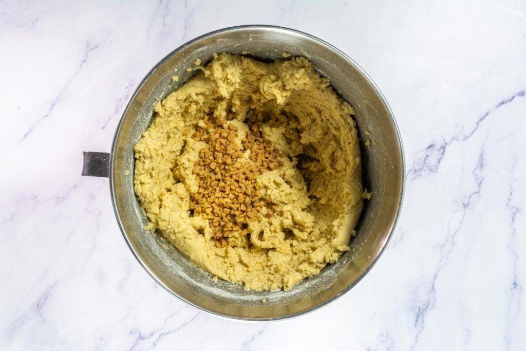 A metal mixing bowl containing cookie dough with toffee bits added in the center, placed on a marble countertop.