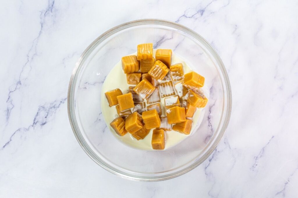 A glass bowl containing caramel candies and sweetened condensed milk on a white marble surface.
