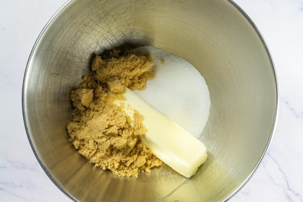 A mixing bowl containing a stick of butter, brown sugar, and granulated white sugar on a white surface.