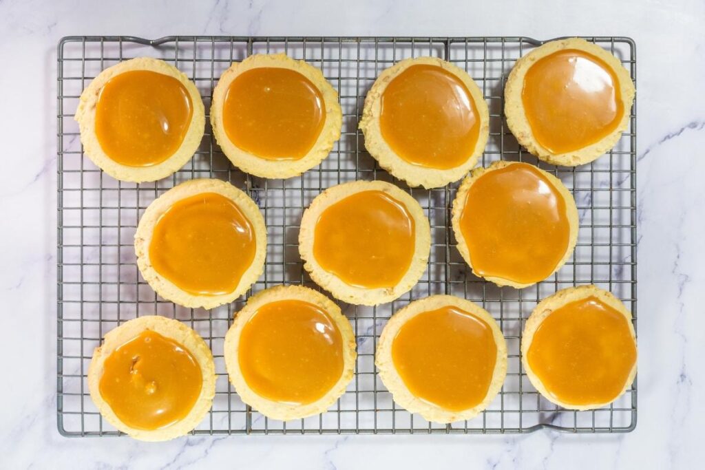 Twelve round cookies with glossy caramel topping are arranged in rows on a cooling rack over a white surface.