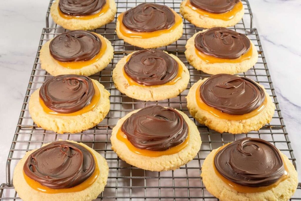 Sugar cookies topped with a layer of caramel and a swirl of chocolate frosting, arranged on a cooling rack.