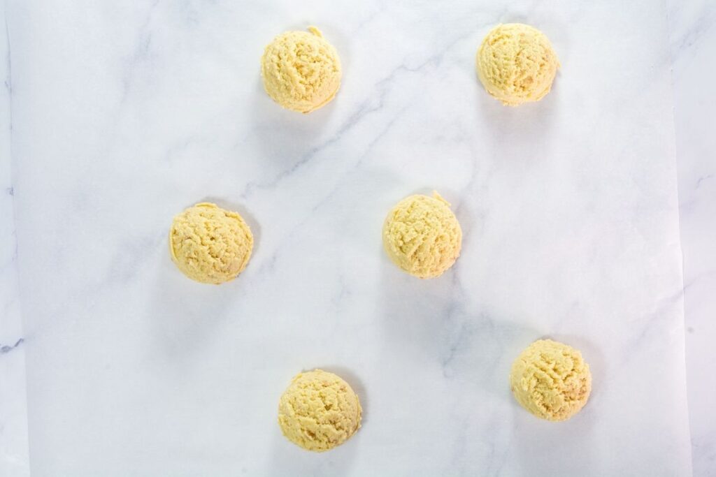Six evenly spaced scoops of cookie dough are arranged on a sheet of parchment paper on a white surface.