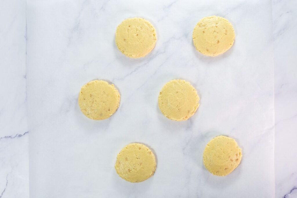 Six round, unbaked cookie dough discs are evenly spaced on a sheet of white parchment paper atop a marble surface.