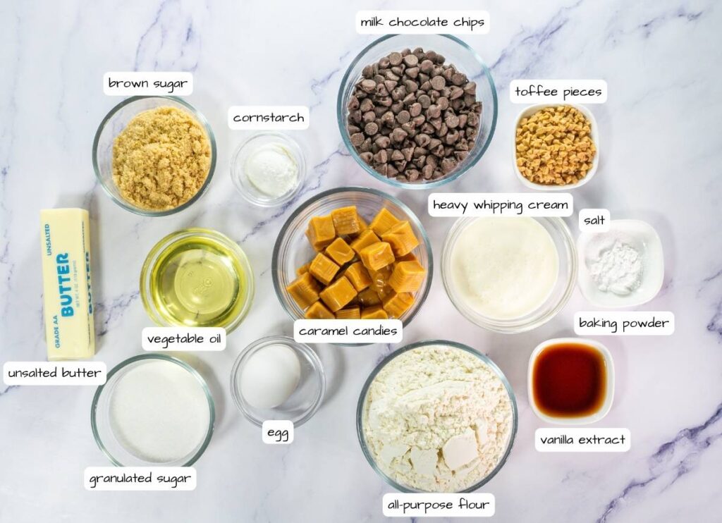 Top-down view of labeled baking ingredients in bowls and containers, including flour, sugars, butter, oil, egg, vanilla, salt, baking powder, cornstarch, caramel candies, toffee pieces, and chocolate chips.