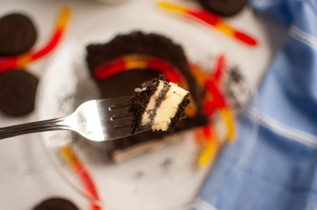 A fork holding a bite of Dirt cake, with gummy worms and crushed chocolate cookies on a plate in the background.