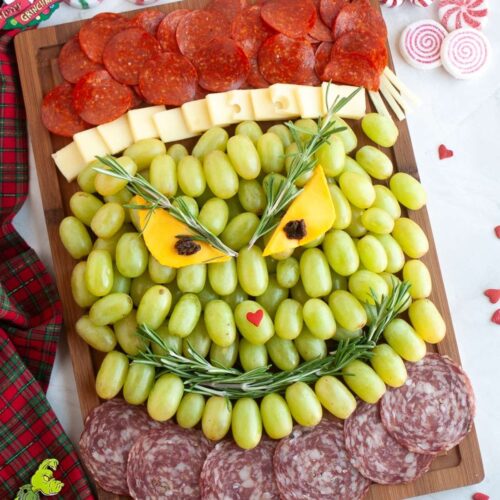 A charcuterie board arranged to resemble the Grinch's face using green grapes, cheese, pepperoni, salami, and herbs on a wooden board with holiday decorations nearby.