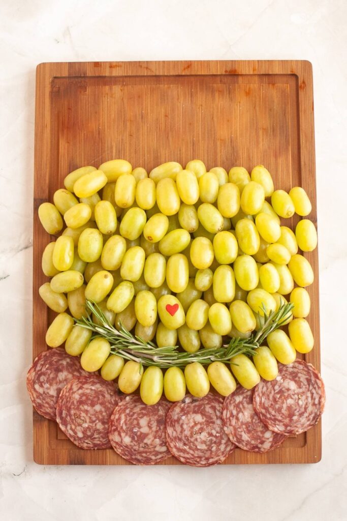 A wooden cutting board with a pile of green grapes, a sprig of rosemary, a small red heart, and slices of salami arranged at the bottom.