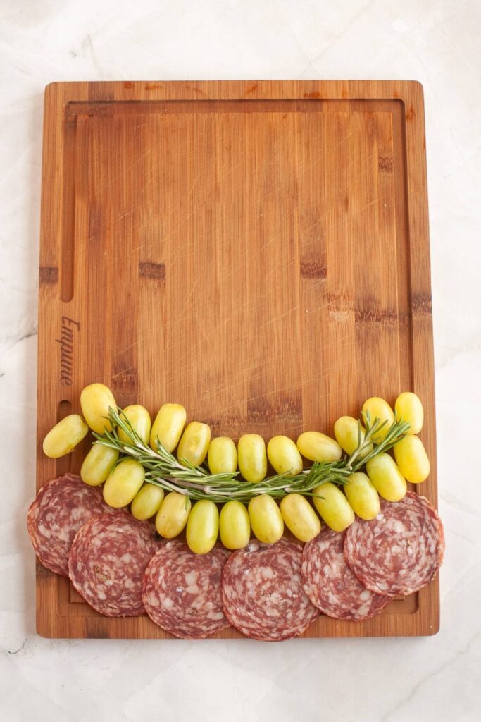 A wooden cutting board with a row of salami slices, a row of green grapes, and a sprig of rosemary arranged along one edge.