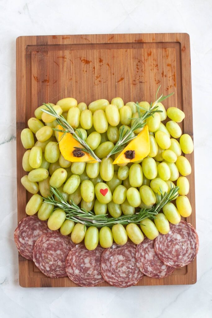 A cutting board with green grapes arranged to look like a face, yellow pepper for eyes, herbs for brows and mouth, and salami slices below resembling a beard.