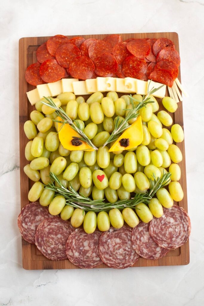 A charcuterie board arranged to resemble the Grinch, with grapes, meat slices, cheese, peppers, rosemary, and a small heart-shaped item on a wooden board.