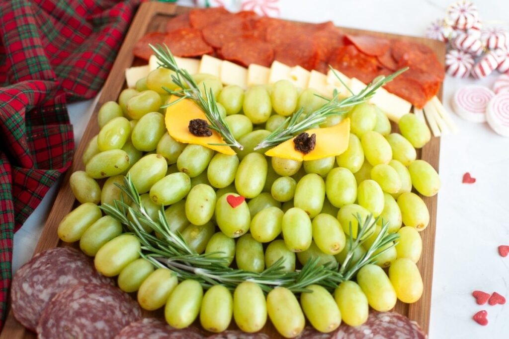 A charcuterie board arranged to resemble a face, with green grapes, cheese, cured meats, and rosemary used for facial features.