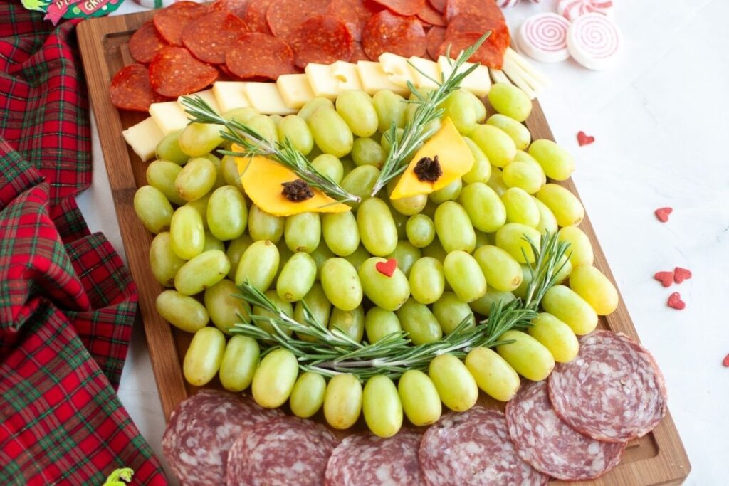 A charcuterie board arranged with green grapes, cheese, meats, and herbs to resemble a face with a mischievous expression.