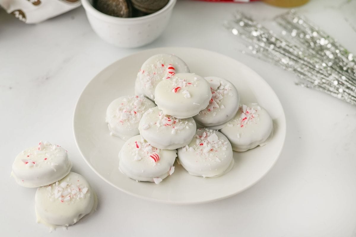 A white plate of Peppermint Crunch Oreos topped with crushed peppermint pieces sits on a white surface.