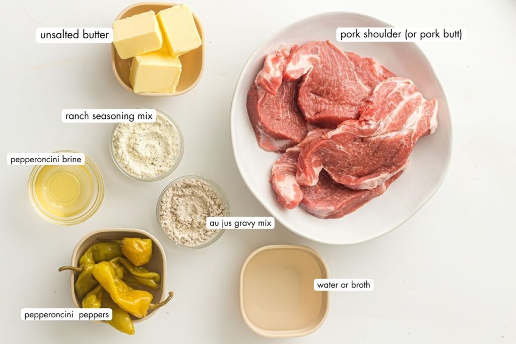 Ingredients for Mississippi pork roast arranged on a white surface: pork shoulder, unsalted butter, ranch seasoning, au jus mix, pepperoncini peppers, pepperoncini brine, and water or broth.