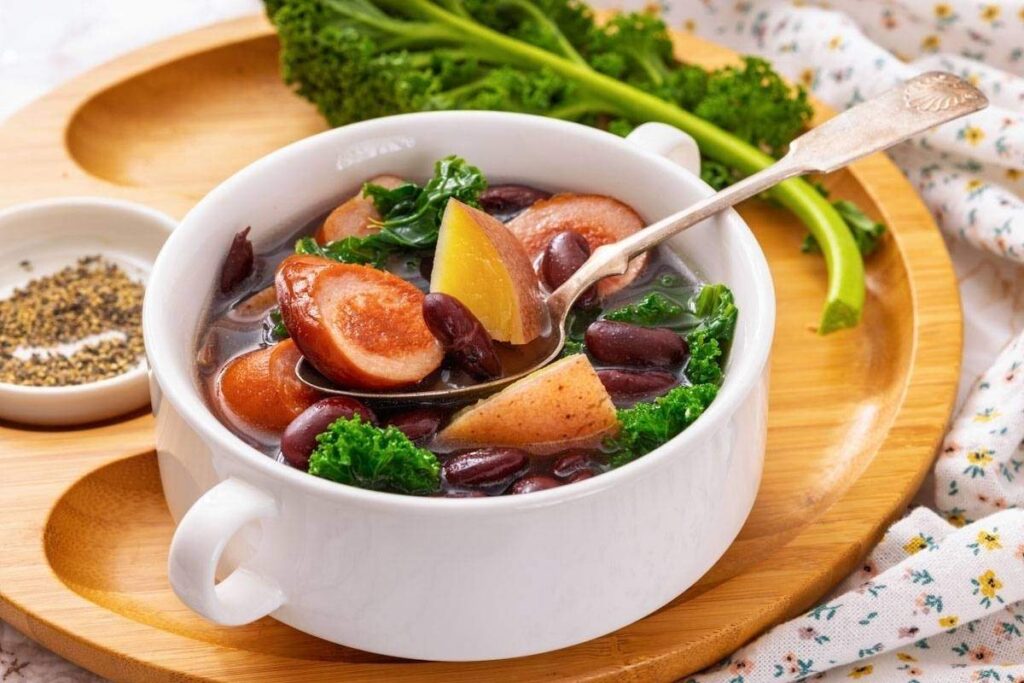 A white bowl of soup containing kale, kidney beans, and sliced sausage sits on a wooden tray, with a spoon in the bowl and black pepper on the side.