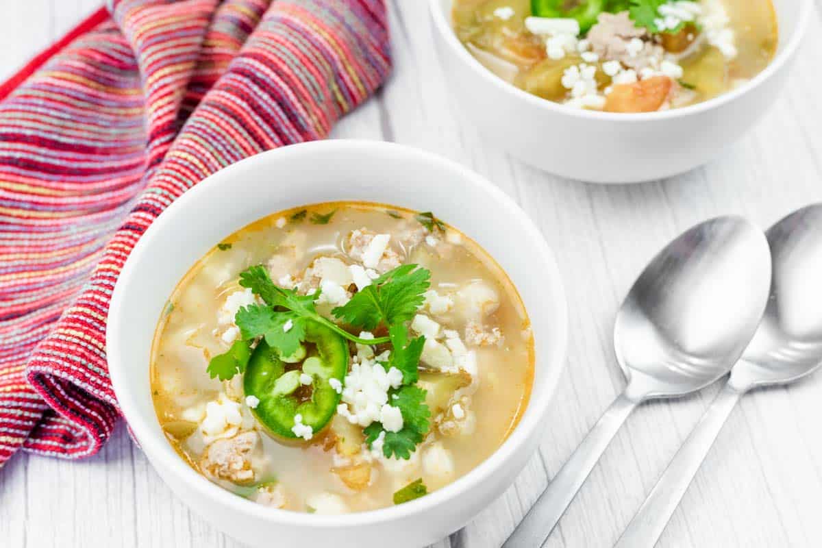 A bowl of Easy Green Pork Posole with white hominy, ground meat, sliced jalapeños, cheese, and cilantro, next to a striped cloth napkin and two metal spoons on a white surface.