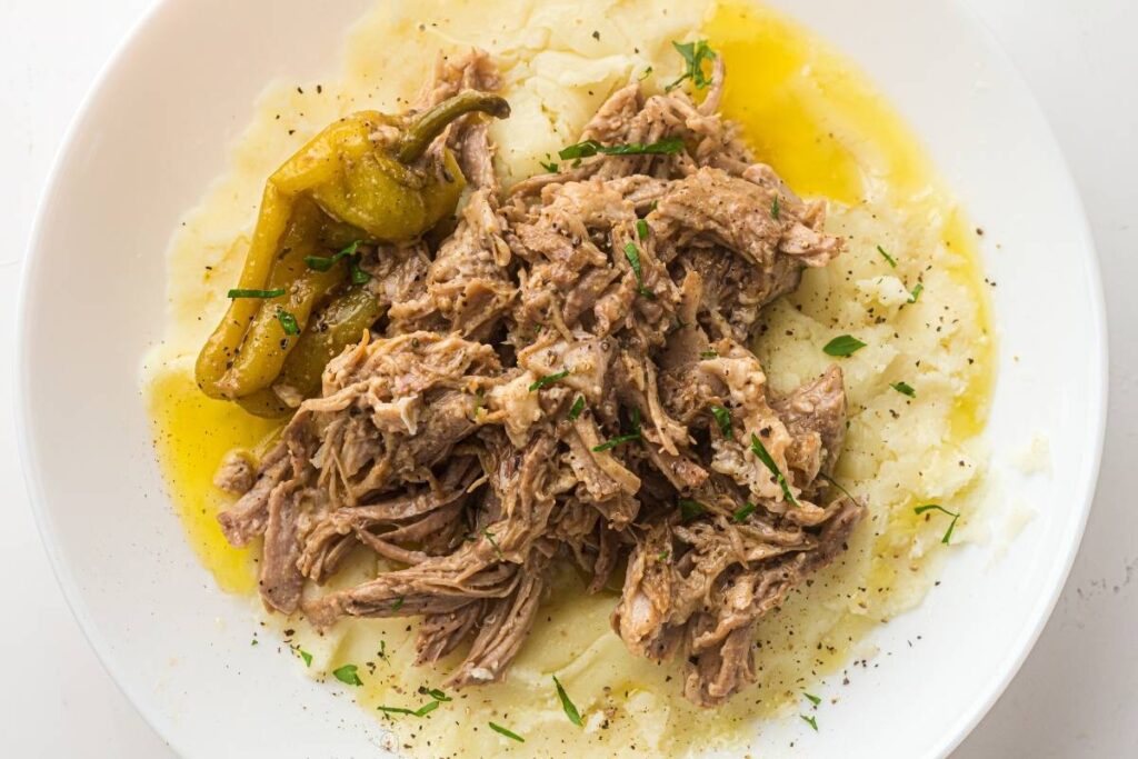 Shredded beef served over mashed potatoes with a whole pepperoncini and garnished with chopped herbs, on a white plate.