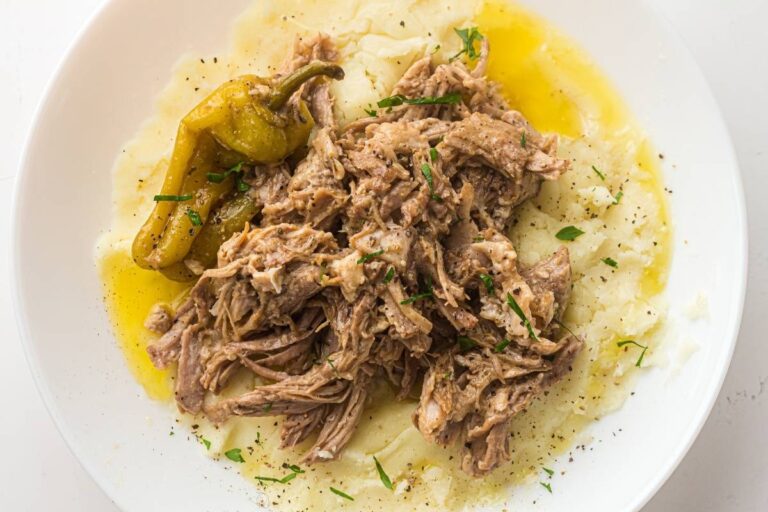 Shredded beef served over mashed potatoes with a whole pepperoncini and garnished with chopped herbs, on a white plate.