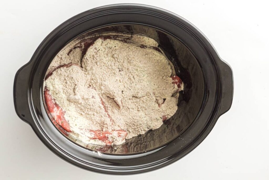 A black slow cooker filled with raw meat covered by a layer of powdered seasoning mix.