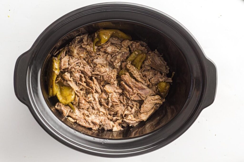 Shredded meat with green peppers in a black slow cooker, viewed from above on a white surface.