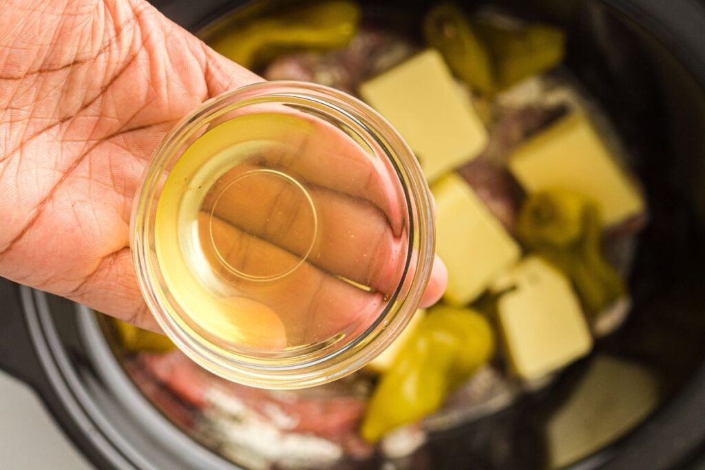 A hand holds a small glass bowl of pepperoncini brine above a slow cooker containing butter, peppers, and other ingredients.