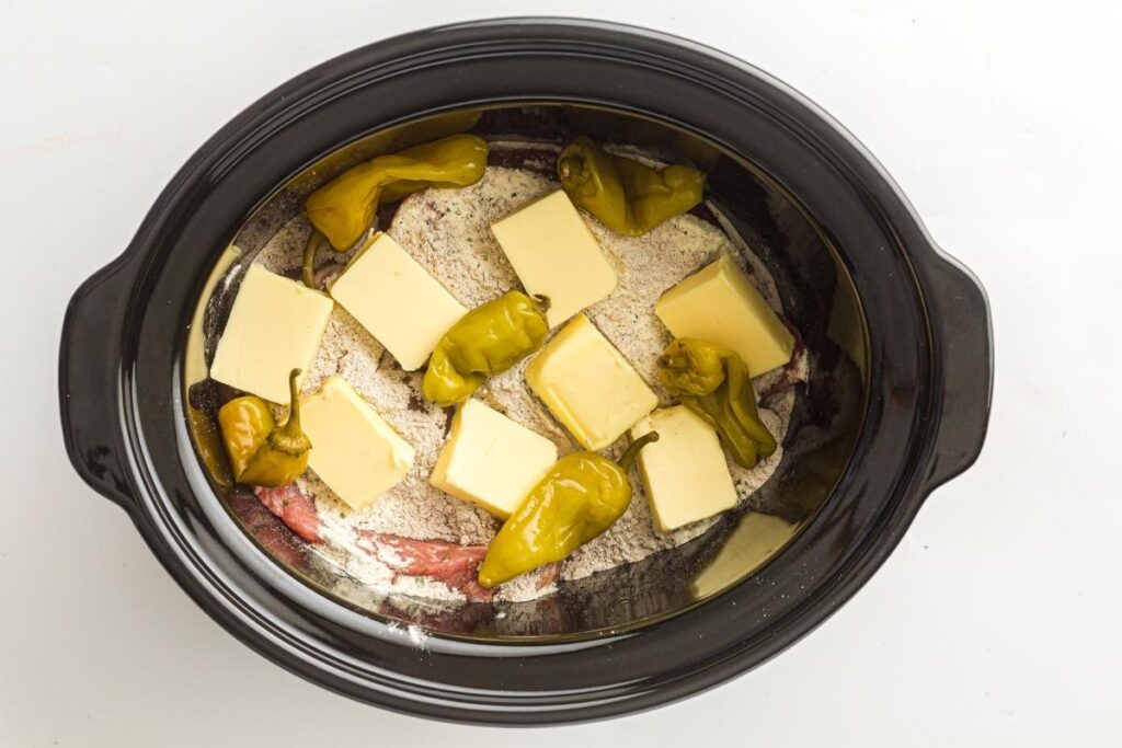 A slow cooker filled with raw beef, dry seasoning mix, sliced butter, and whole pepperoncini peppers, ready to be cooked.