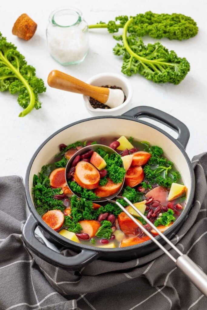 A pot of vegetable soup with carrots, potatoes, kidney beans, and kale sits on a gray cloth, with fresh kale, salt, and pepper nearby.