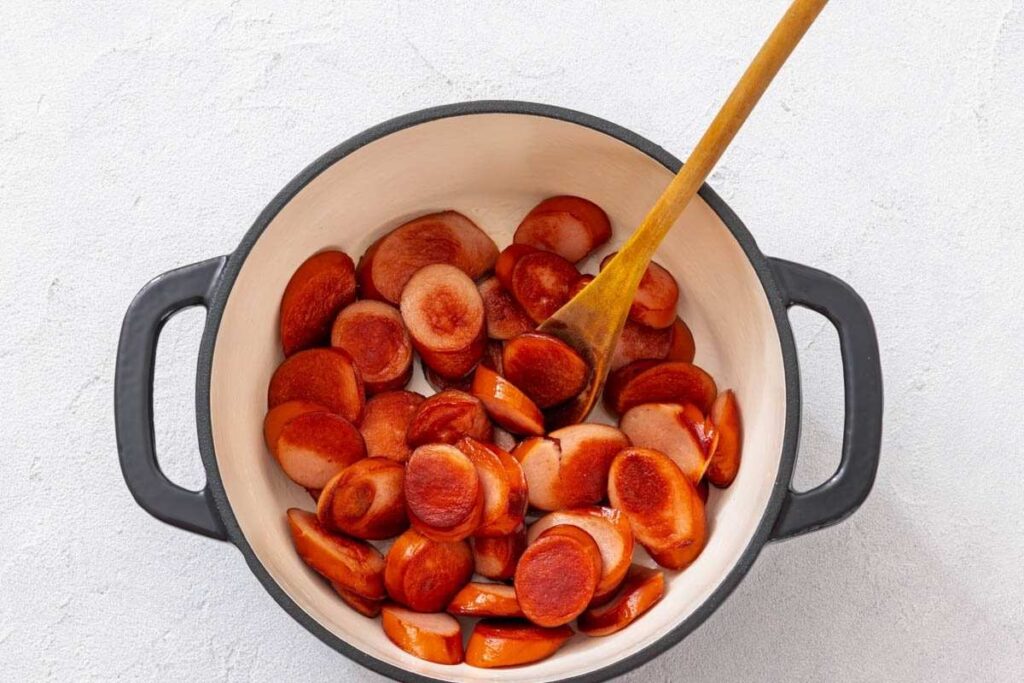 Sliced sausage pieces are being cooked in a white pot with a black rim, stirred by a wooden spoon.